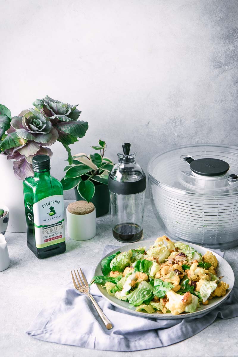 a salad, salad spinner, salad dressing shaker, bottle of olive oil, and vase with flowers on a white table