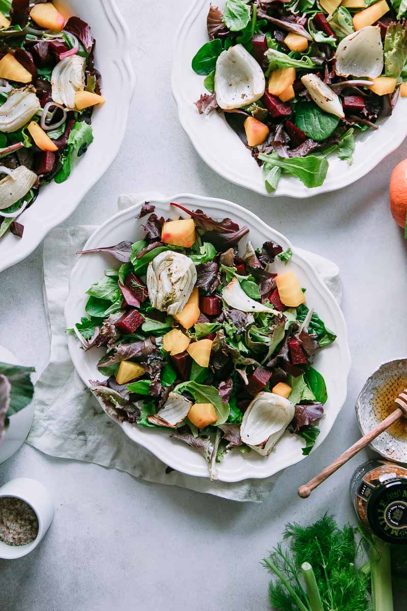 Three salads with beets on a white table with a honey pot and dipper, flowers, and an orange.