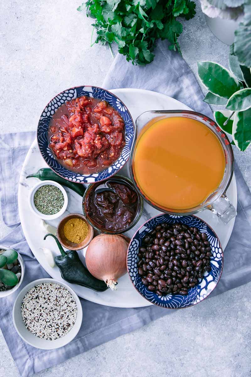 a round cutting board with vegetable broth, black beans, fire roasted tomatoes, peppers, and spices in small bowls on a white table