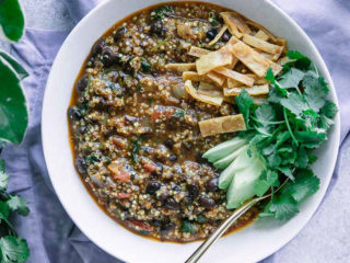 a white bowl of black bean chili topped with avocado, cilantro, and tortillas strips on a white table with a blue napkin