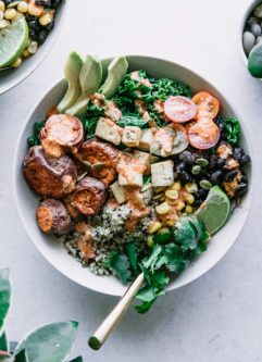 quinoa, black beans, kale, corn, tomatoes, roasted sweet potatoes, sliced avocado, chipotle lime tahini sauce, a lime wedge, and cilantro in a white bowl on a white countertop with two green plants