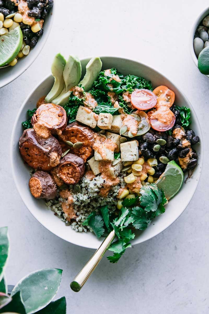 quinoa, black beans, kale, roasted sweet potatoes, sliced avocado, chipotle lime tahini sauce, a lime wedge, and cilantro in a white bowl on a white countertop with lime wedges