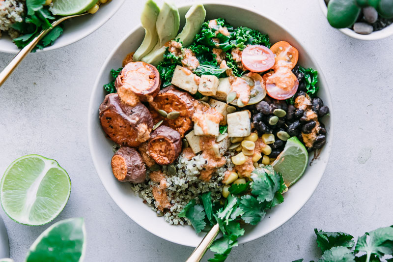 a white bowl with quinoa, black beans, tofu, sweet potatoes, and a creamy red chipotle chili lime sauce