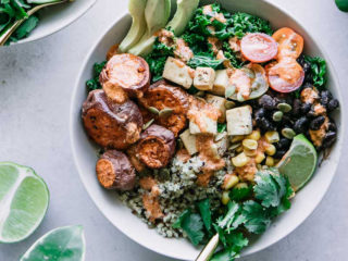 quinoa, black beans, corn, kale, tomatoes, roasted sweet potatoes, sliced avocado, a lime wedge, and cilantro in a white bowl on a white countertop with lime wedges and cilantro leaves, and the words "chipotle lime black bean bowl" in black writing with green and yellow highlight