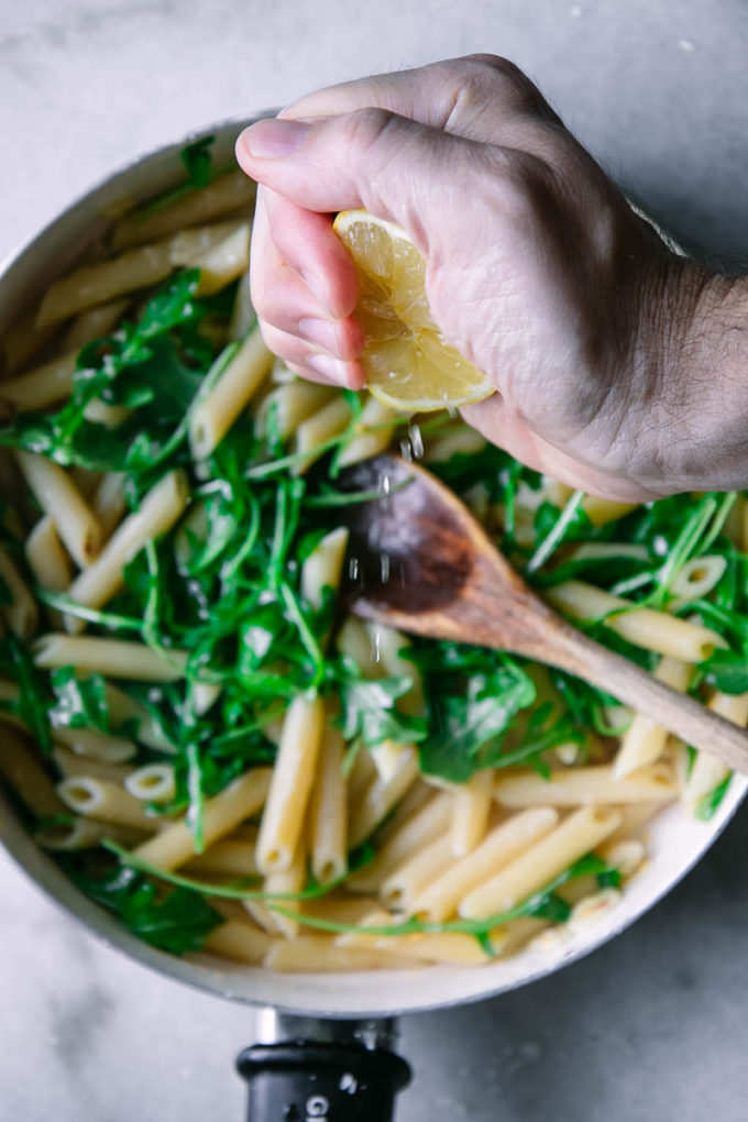 a hand squeezing lemon into a pan of pasta with arugula