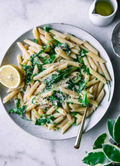 penne, arugula, and lemon pasta on a white plate with a gold fork on a marble countertop with a green plant
