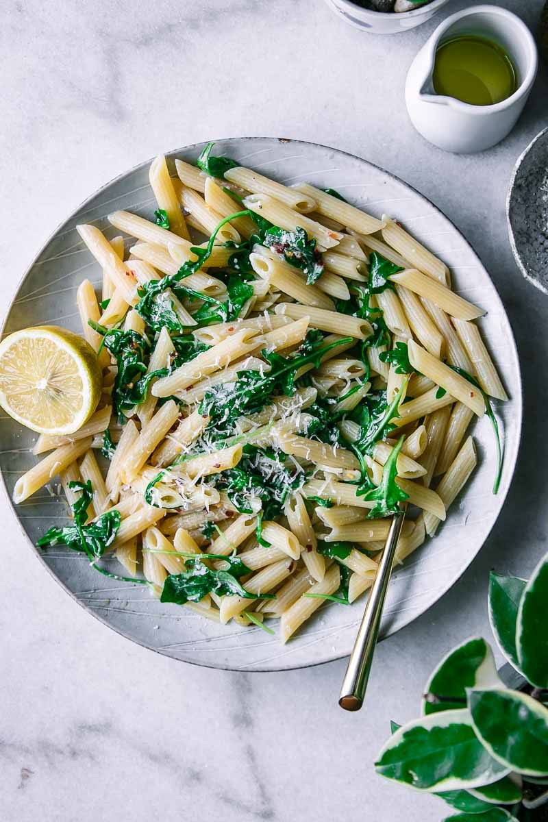 penne, arugula, and lemon pasta on a white plate with a gold fork on a marble countertop with a green plant