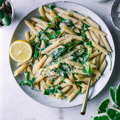 a plate of penne pasta with spinach and a slice of lemon on a white table with a bowl of parmesan cheese