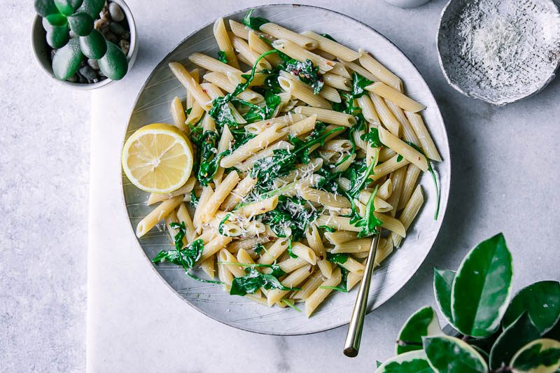 a plate of penne pasta with spinach and a slice of lemon on a white table with a bowl of parmesan cheese