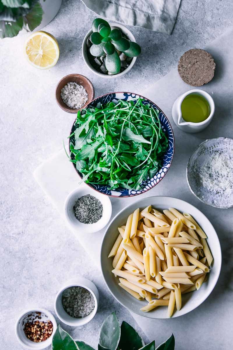 bowls of penne, arugula, chili flakes, olive oil, sea salt, and parmesan cheese on a white table with a green plant