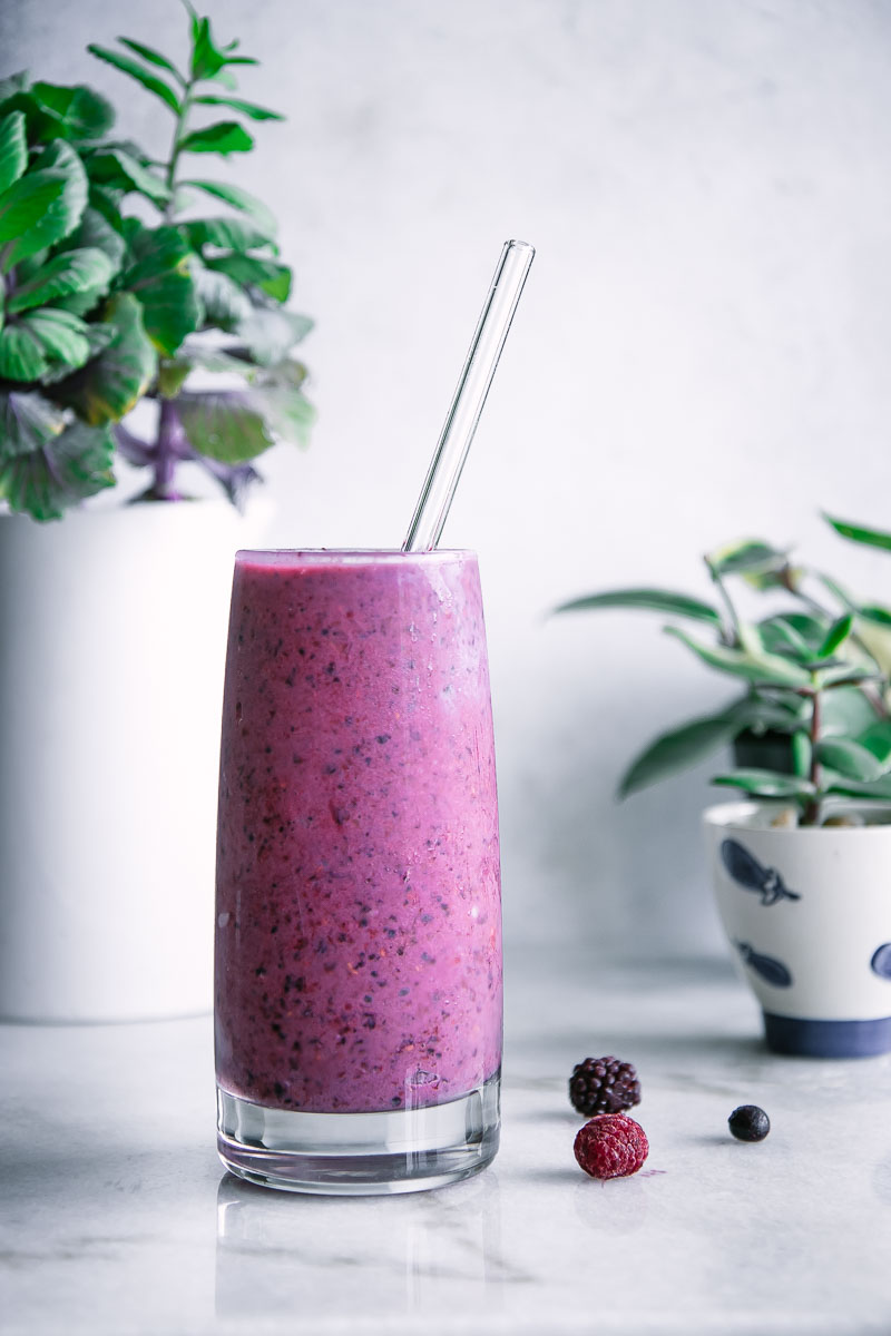 a tall glass with a purple smoothie and a glass straw on a white table with green plants