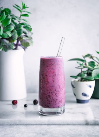 a tall glass with a bright smoothie with a glass straw with blueberries, blackberries, and raspberries and bananas on a marble table