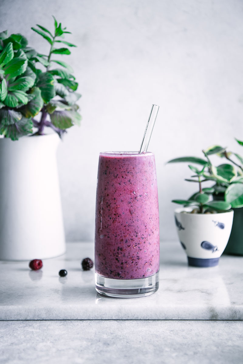 a tall glass with a bright smoothie with a glass straw with blueberries, blackberries, and raspberries and bananas on a marble table