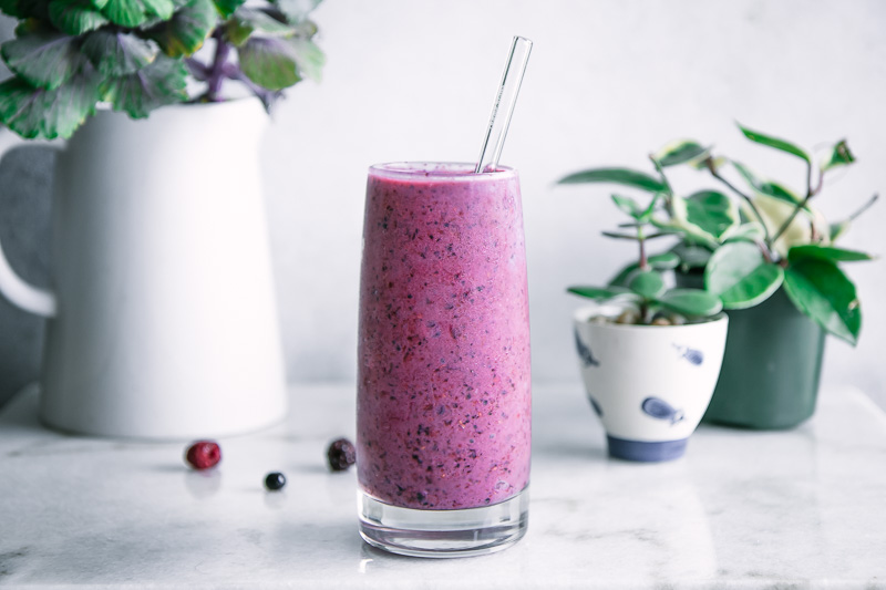 a breakfast smoothie with three berries, bananas, protein powder, and plant-based milk on a white table with green plants in the background.
