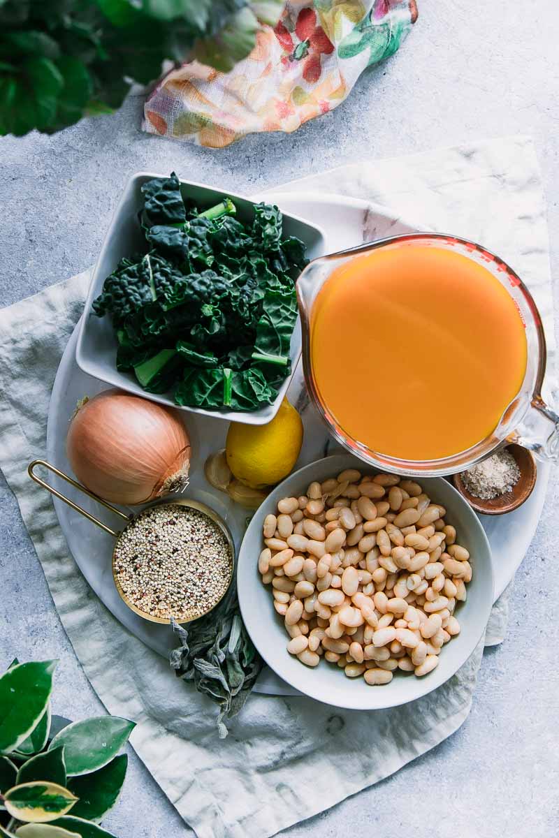 A round white plate with vegetable broth, a bowl of white beans, a bowl of quinoa, a whole onion, a whole lemon, and a cup of kale on a white table