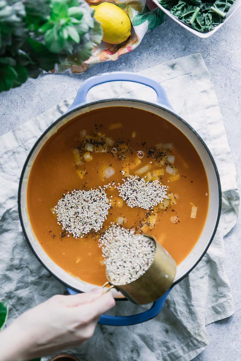 a hand pouring a bowl of quinoa into a large soup pot with broth