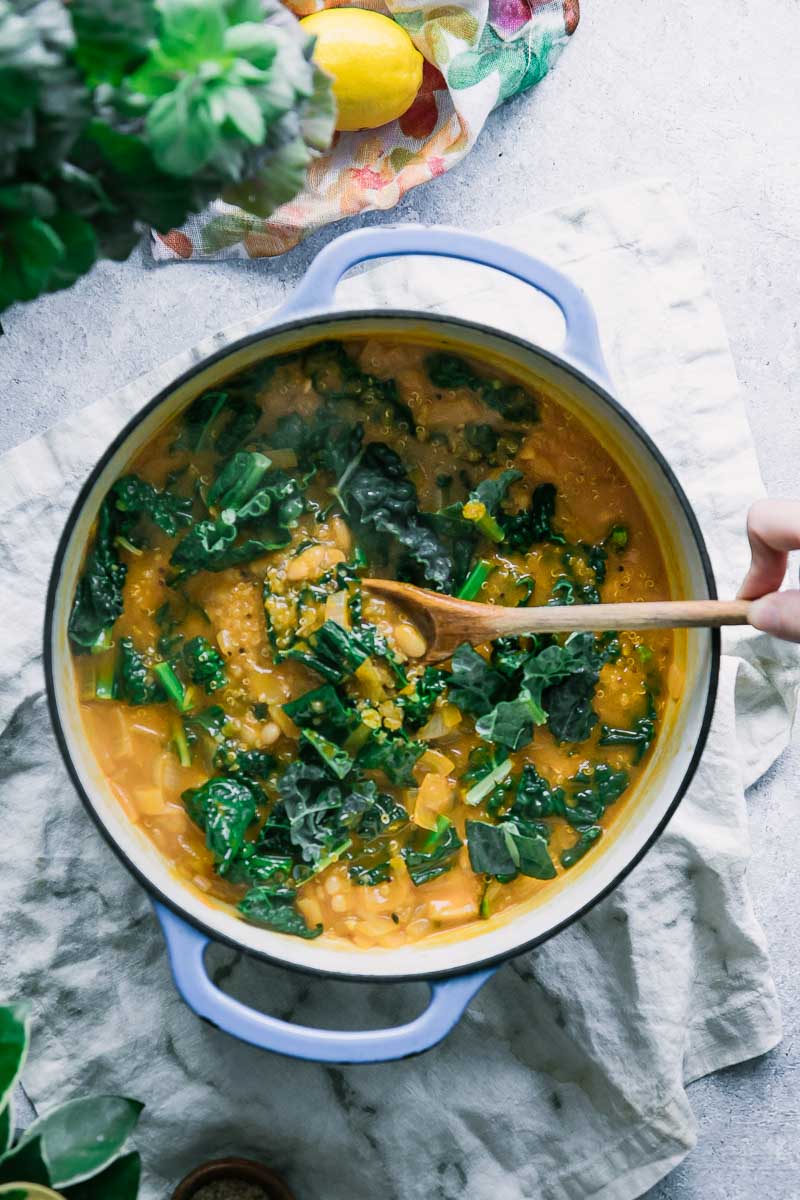 A hand stirring a soup pot with kale with a wooden spoon on a white table