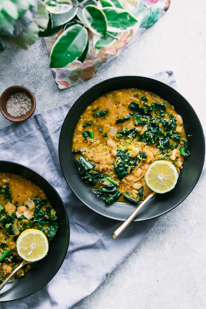 two black bowls with yellow soup with beans and quinoa and kale on a white table with a blue napkin and a wooden bowl of salt and a green plant