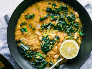 a black bowl with an orange soup with white beans, kale, and a sliced lemon on a blue table with a graphic that says "white bean kale quinoa chili" in black writing