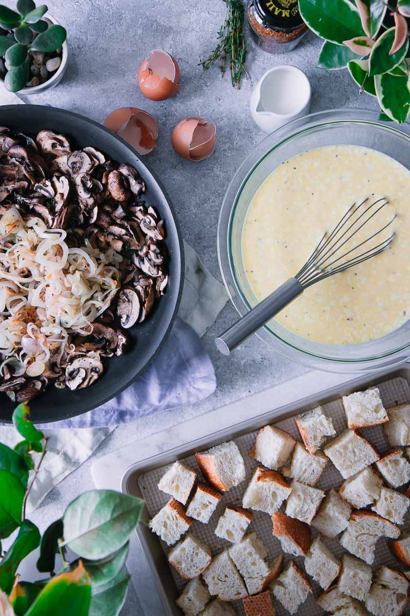 a sheetpan with toasted bread, a pan with cooked mushrooms and shallots, and a bowl with eggs and cream with a whisk, all on a blue table with napkins