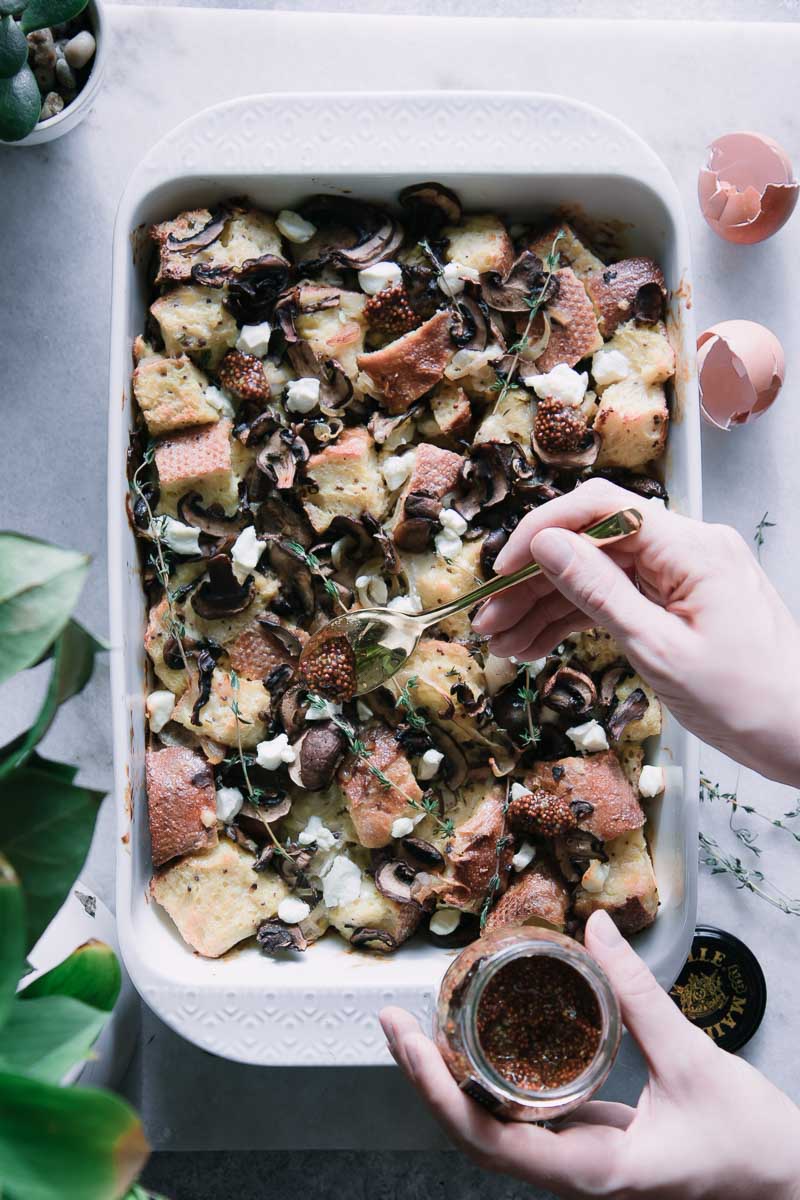 a white baking dish with strata made from sourdough and mushrooms, with a hand drizzling mustard sauce on top