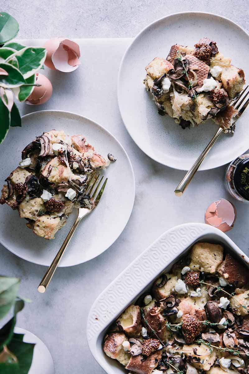 A baked mushrooms sourdough breakfast strata in a white baking dish with two plate with gold forks