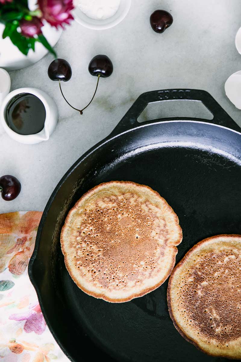 two hoecakes on a black cast iron skillet with cherries and flowers on a white table