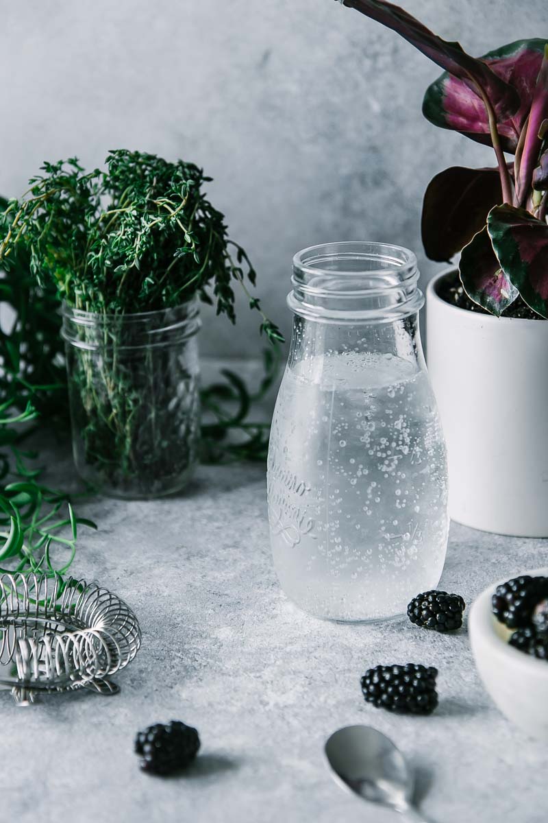 a caraffe of sparkling water, a bowl of blackberries, and a jar of fresh thyme on a blue table