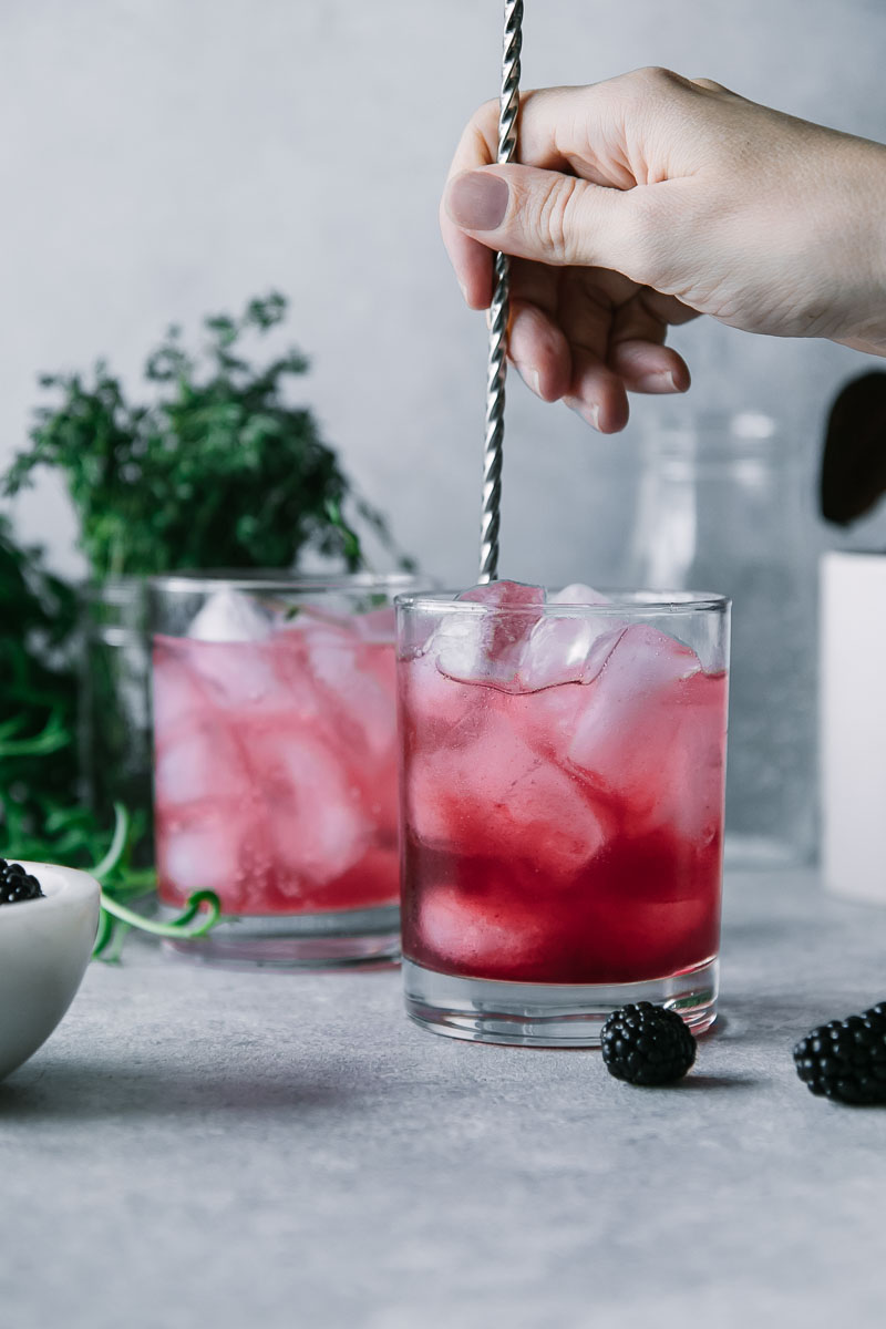 a hand mixing a red cocktail with a mixing spoon on a blue table