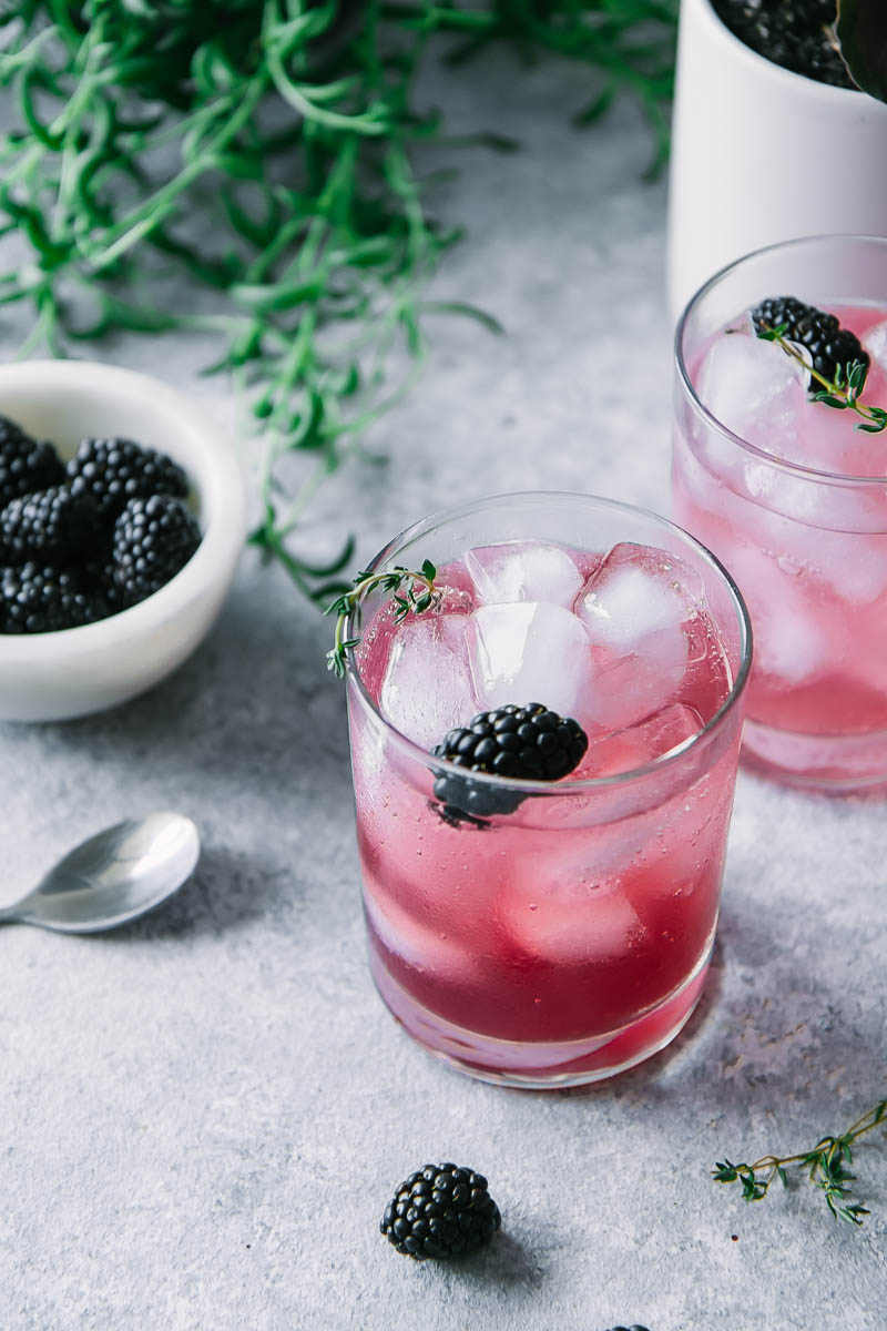 two glasses of sparkling water with blackberry and thyme over ice on a table with a bowl of blackberries