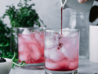 a hand pouring blackberry juice into a glass of sparkling water with the words "Blackberry Thyme Spritzer" in black writing