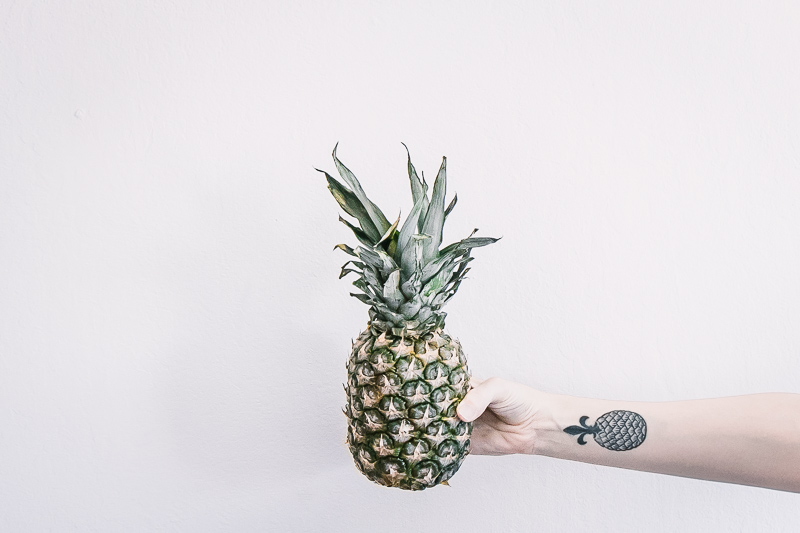 a horizontal ratio photo of a hand with a pineapple tattoo holding a pineapple with a white background