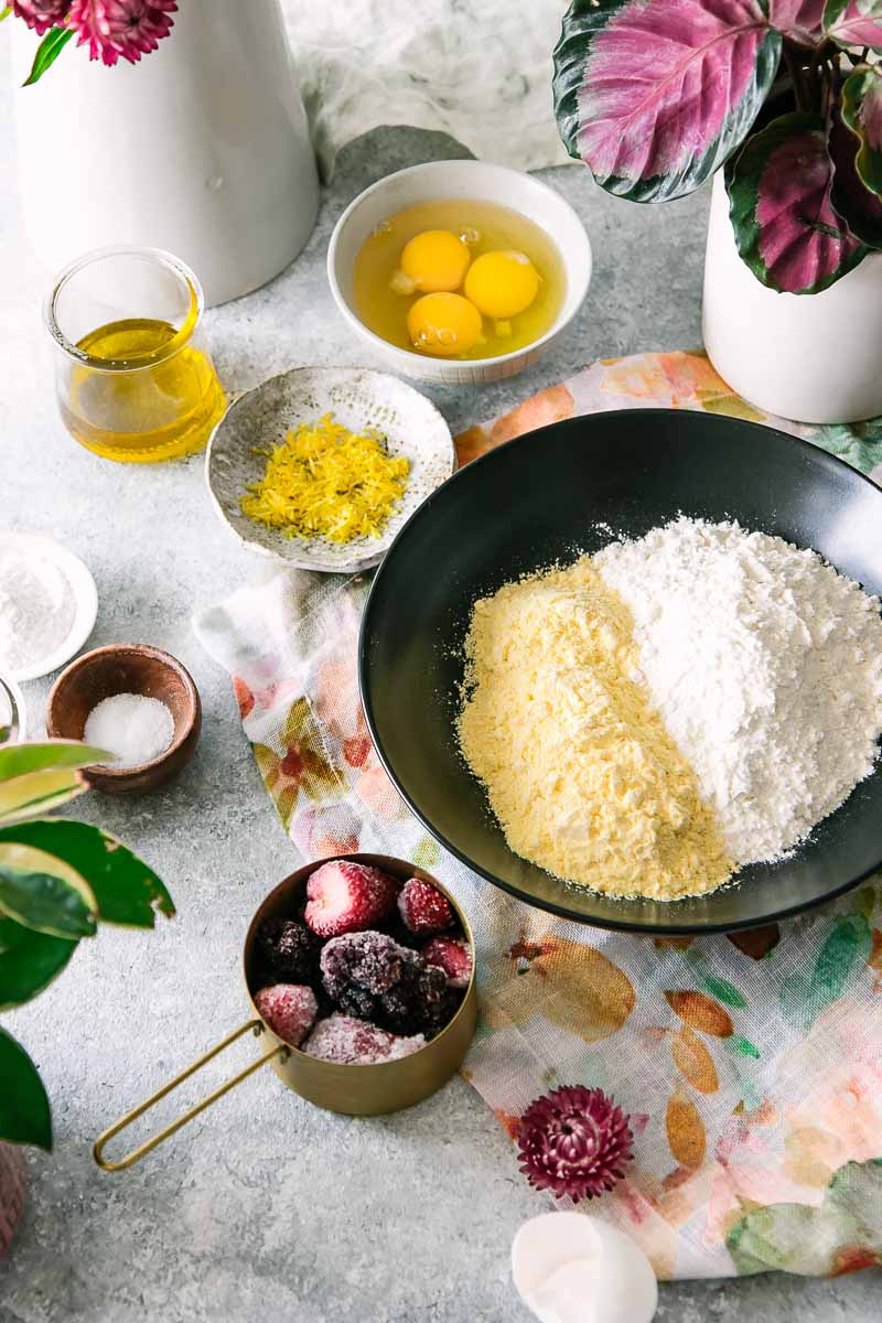a bowl of flour, cornmeal, eggs, lemon zest, oil, salt, and frozen berries on a blue table