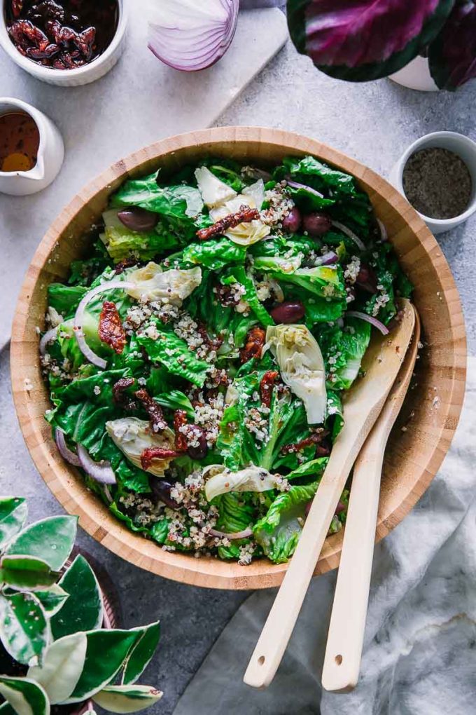 a wooden salad bowl with romaine lettuce, red onion, kalamata olives, and quinoa on a white table.