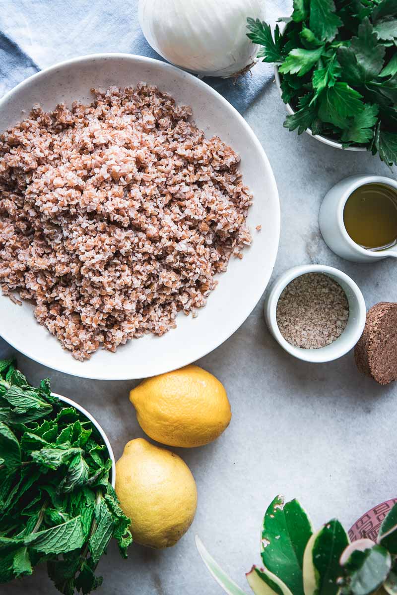 bowls of bulgur, parsley, mint, and lemons, salt, and onion on a white marble table