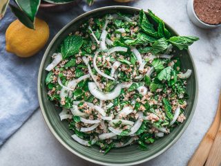 a green bowl with traditional tabbouleh with bulgur, onion, parsley, and mint on a white table with a blue napkin and lemons