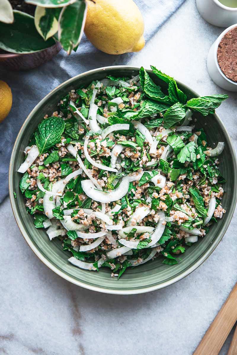 a green bowl with bulgur tabbouleh, mint, and parsley and two lemons on a white table