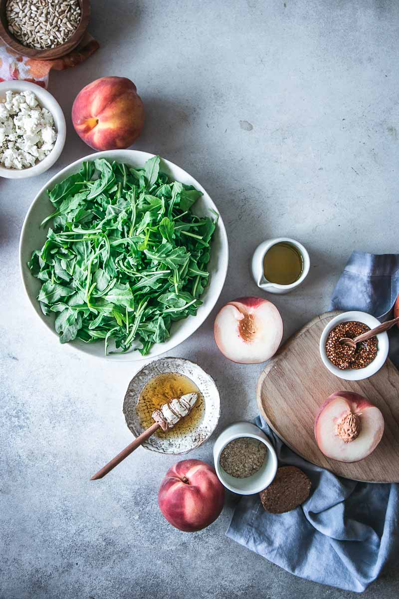 a flatlay photo of summer grilled peach salad ingredients in bowls on a blue table, including arugula, peaches, honey, sunflower seeds, goat cheese, dijon mustard, olive oil, and salt