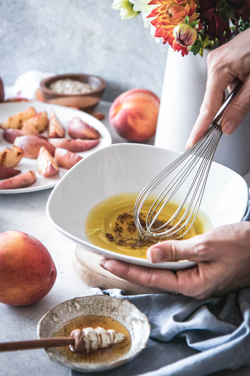 a hand mixing olive oil, apple cider vinegar, mustard, and honey in a white bowl with a whisk on a table with a salad and peaches in the background