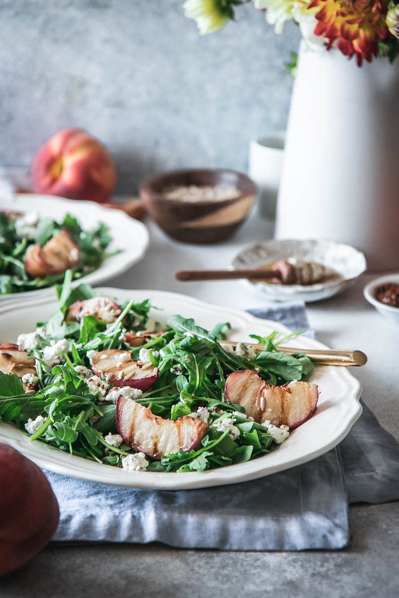 a white plate with arugula and grill peach salad on a blue table with a vase of orange flowers and peaches