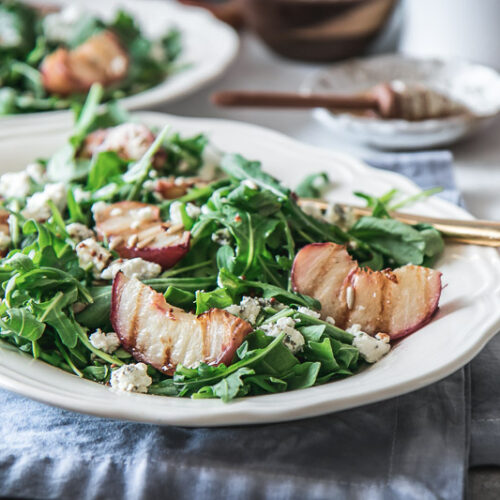 a white plate with a salad with grilled peaches, arugula, and goat cheese on a white table with a peach and a bowl of honey