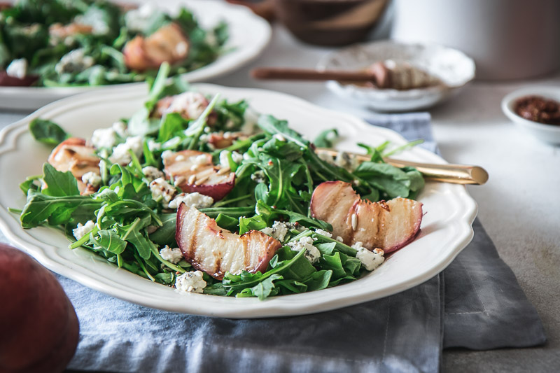 a white plate with a salad with grilled peaches, arugula, and goat cheese on a white table with a peach and a bowl of honey