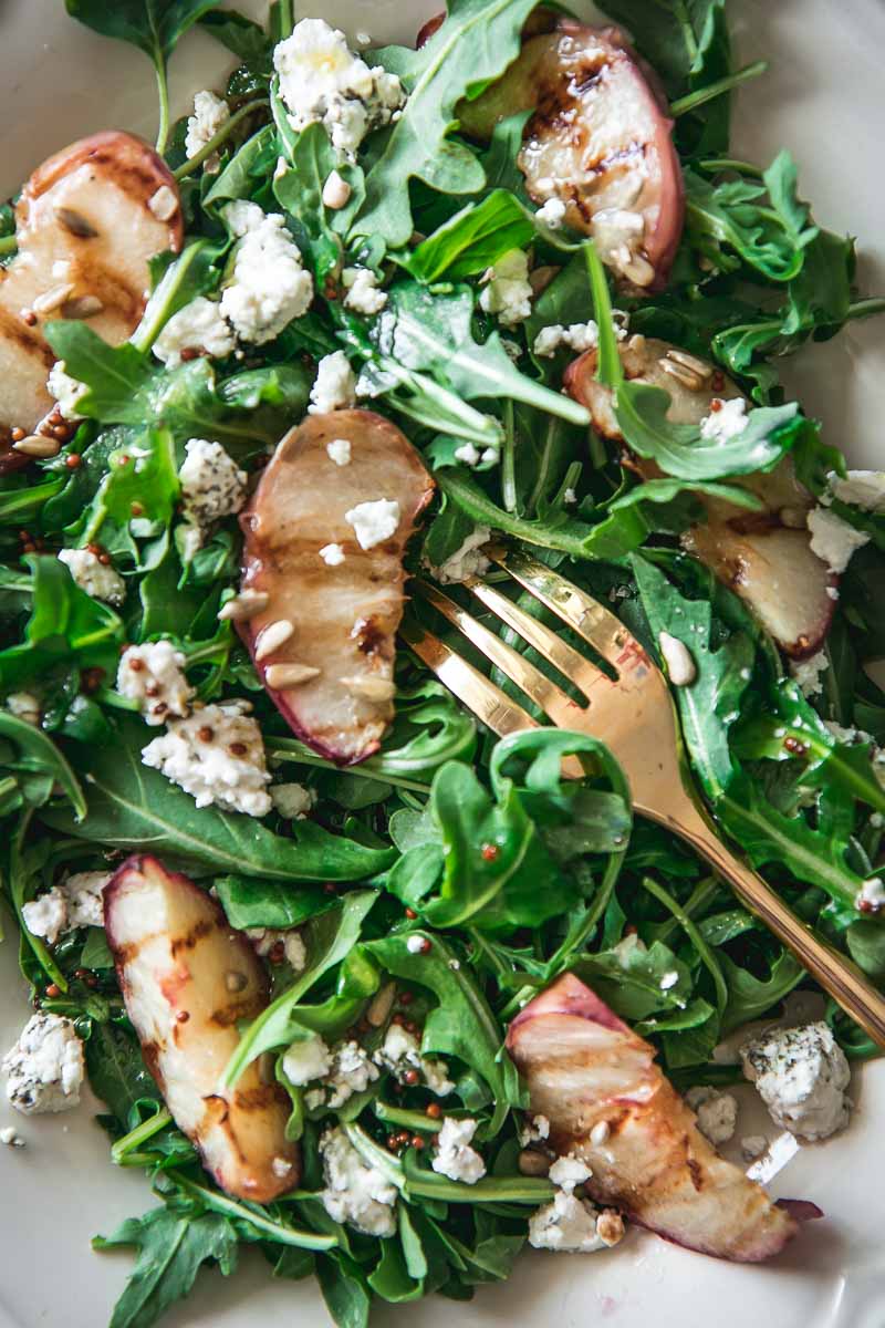 a close up shot of an arugula, grilled peach, goat cheese, and sunflower salad with a gold fork