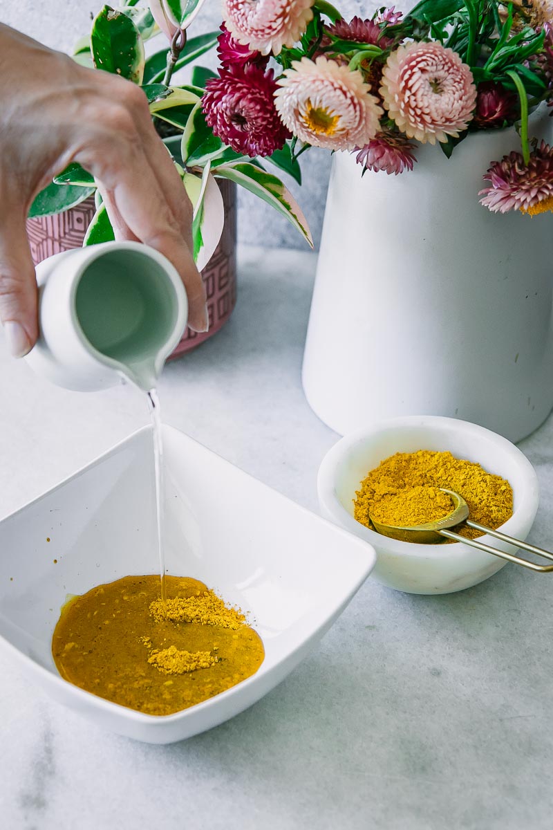 a hand pouring water from a pitcher into a bowl of turmeric and golden milk spices in a white bowl