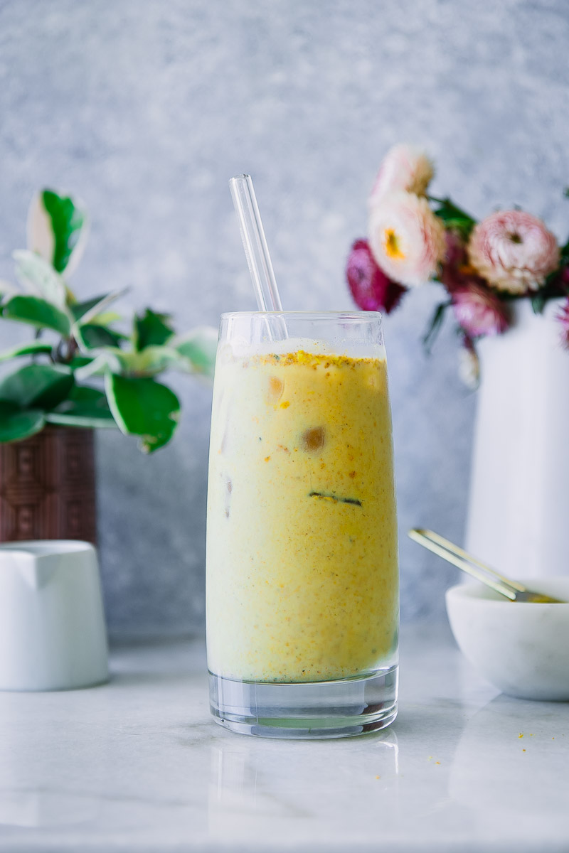 a tall glass with ice filled with a golden milk latte with yellow turmeric on a white table with pink flowers