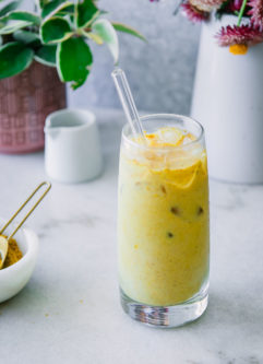 a glass of yellow golden milk with ice on a blue table with turmeric spice, flowers, and vegan milk in the background