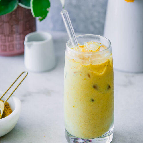 a glass of yellow golden milk with ice on a blue table with turmeric spice, flowers, and vegan milk in the background
