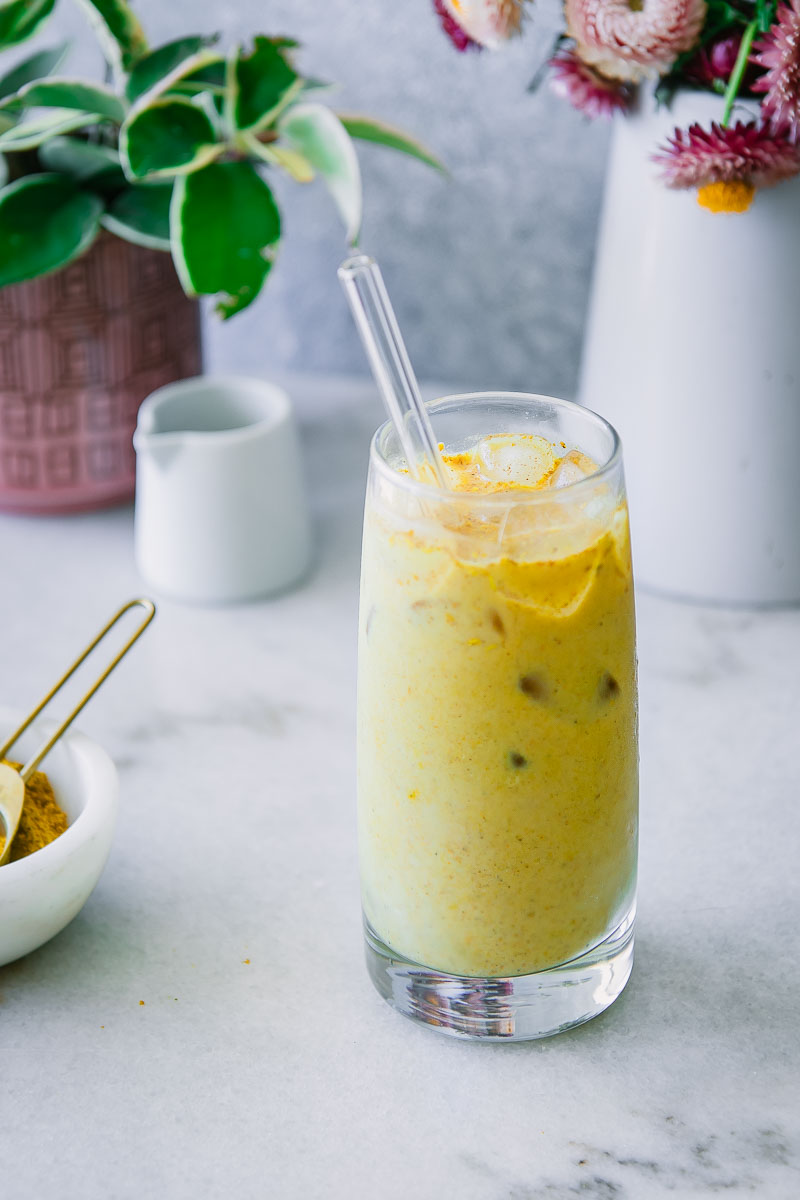 a glass of yellow golden milk with ice on a blue table with turmeric spice, flowers, and vegan milk in the background