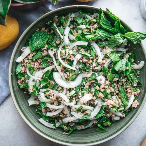 a green bowl with traditional tabbouleh with bulgur, onion, parsley, and mint on a white table with a blue napkin and lemons