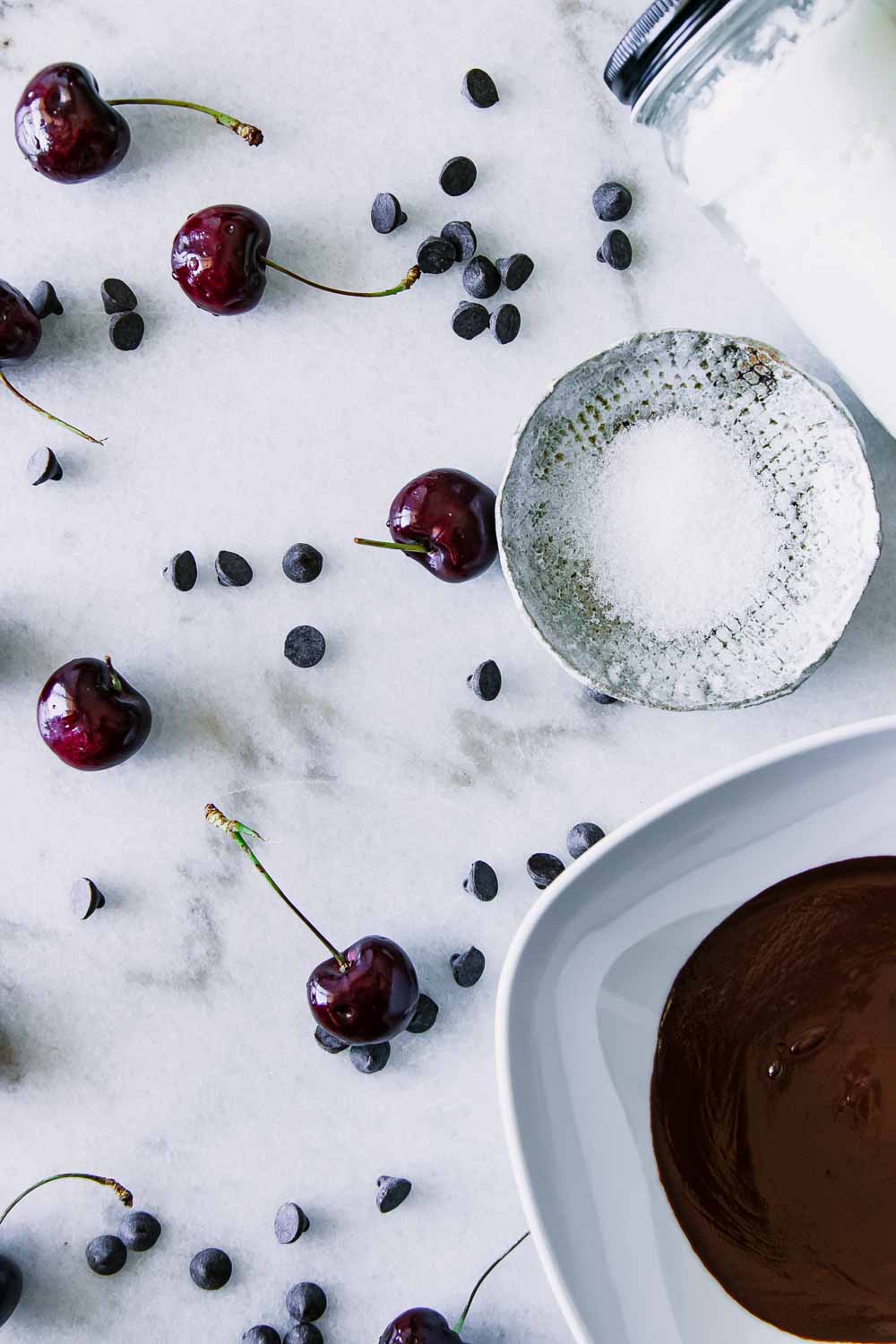 black cherries, dark chocolate chips, a bowl of sea salt, and a bowl of melted chocolate on a white marble table